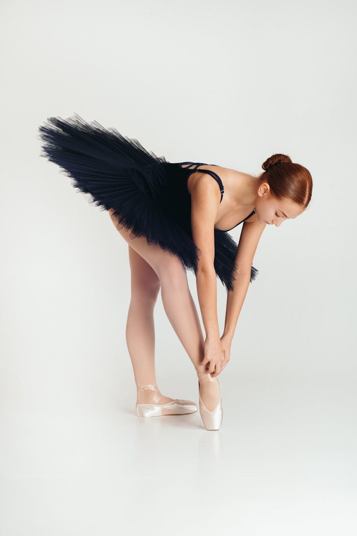 A graceful ballerina in a black tutu adjusts her shoes against a simple white background.